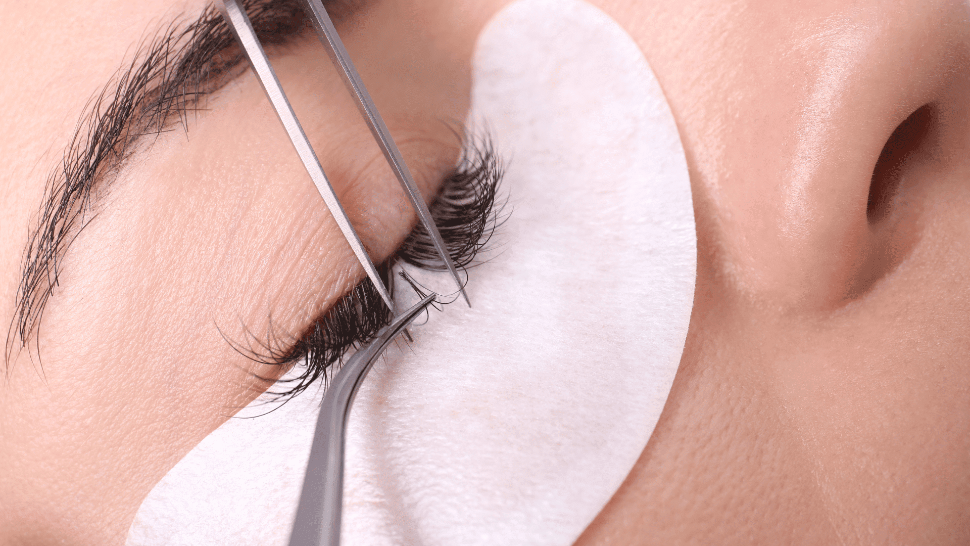 Close-up of a person getting eyelash extensions applied with tweezers onto a cushioned eye pad.