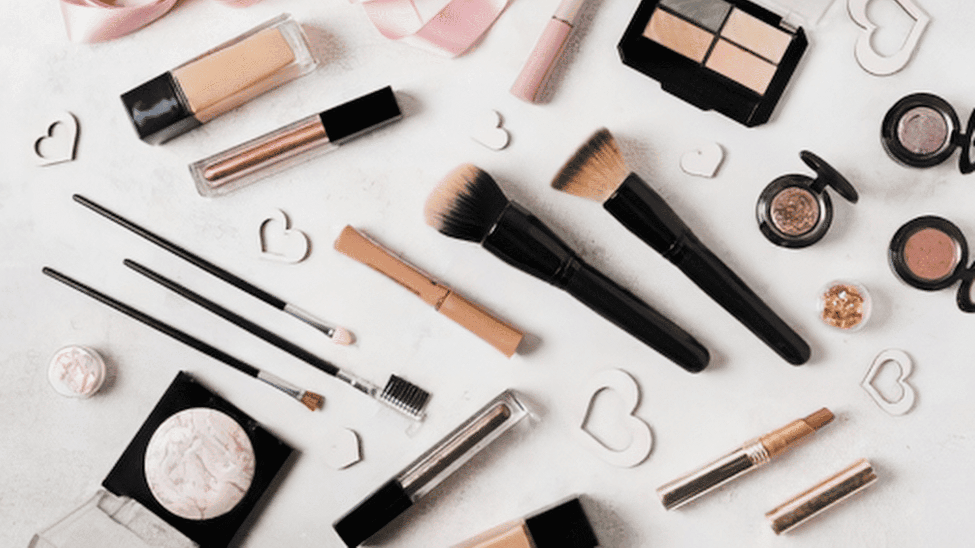 Makeup items and brushes arranged on a white surface with small heart decorations.